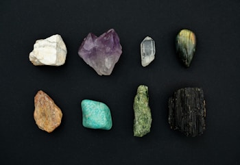 A collection of eight distinct mineral stones arranged in two rows against a dark background. The stones exhibit a variety of colors and textures, ranging from white to deep green and black, with some displaying a crystalline structure.