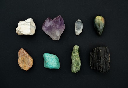 A collection of eight distinct mineral stones arranged in two rows against a dark background. The stones exhibit a variety of colors and textures, ranging from white to deep green and black, with some displaying a crystalline structure.