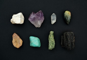 A collection of eight distinct mineral stones arranged in two rows against a dark background. The stones exhibit a variety of colors and textures, ranging from white to deep green and black, with some displaying a crystalline structure.