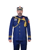 A person is wearing a formal military or naval uniform with a white and blue cap. The uniform is primarily navy blue, adorned with various gold accents including epaulettes, buttons, and decorative cords. There are multiple badges and insignia on the chest and arms, and a flag patch on the right sleeve.