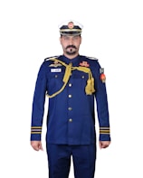 A person is wearing a formal military or naval uniform with a white and blue cap. The uniform is primarily navy blue, adorned with various gold accents including epaulettes, buttons, and decorative cords. There are multiple badges and insignia on the chest and arms, and a flag patch on the right sleeve.