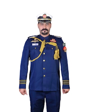 A person is wearing a formal military or naval uniform with a white and blue cap. The uniform is primarily navy blue, adorned with various gold accents including epaulettes, buttons, and decorative cords. There are multiple badges and insignia on the chest and arms, and a flag patch on the right sleeve.