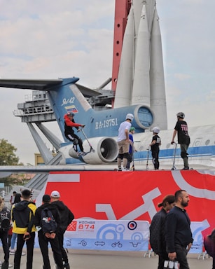 A group of people are engaged in an outdoor activity involving scooters. They are positioned on a platform in front of a large aircraft, and a rocket model is visible in the background. Several onlookers are standing nearby, observing the activity. The scene has a mix of vibrant and neutral colors, with an urban setting.