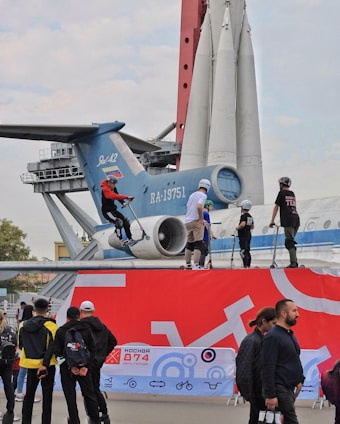 A group of people are engaged in an outdoor activity involving scooters. They are positioned on a platform in front of a large aircraft, and a rocket model is visible in the background. Several onlookers are standing nearby, observing the activity. The scene has a mix of vibrant and neutral colors, with an urban setting.