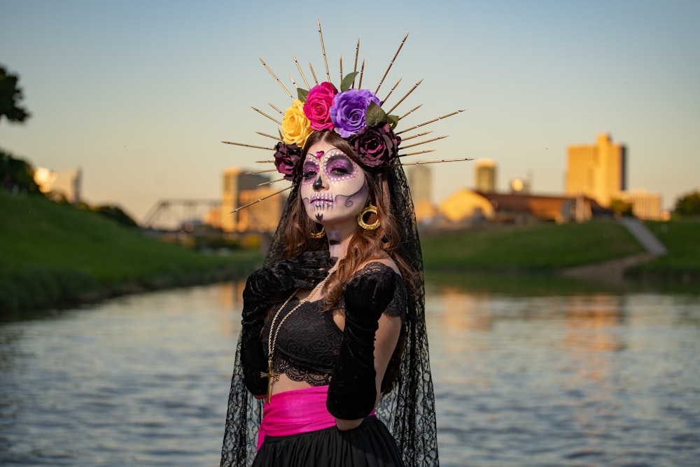 a woman in a skeleton makeup and headdress stands near a body of water