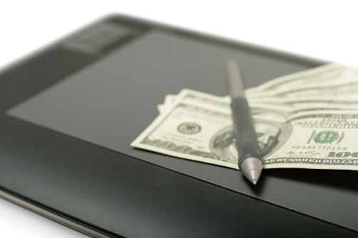 A stack of hundred-dollar bills is placed on top of a digital tablet. A stylus pen rests on the bills, suggesting a context of digital finance or business.