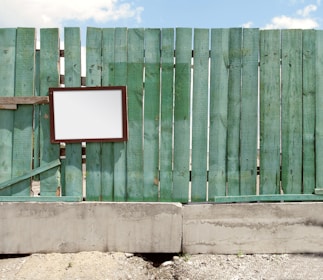 A weathered green wooden fence with vertical planks stands against a clear blue sky. A blank, rectangular sign with a dark frame is attached to the fence. The base of the fence is made of concrete, and the ground is covered in gravel.