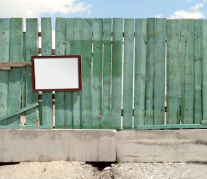 Close-up of a teal and gray campaign sign resting against an old wooden fence on a neighborhood street.