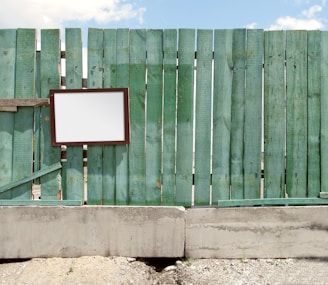 A weathered green wooden fence with vertical planks stands against a clear blue sky. A blank, rectangular sign with a dark frame is attached to the fence. The base of the fence is made of concrete, and the ground is covered in gravel.