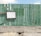A weathered green wooden fence with vertical planks stands against a clear blue sky. A blank, rectangular sign with a dark frame is attached to the fence. The base of the fence is made of concrete, and the ground is covered in gravel.