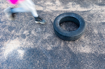 A person is running or jumping, with only their lower half visible in motion blur. They are wearing black sports shoes and white leggings with a pink detail. An old tire is lying on a textured ground surface, possibly for training or exercise purposes.