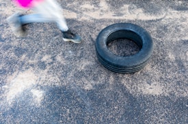 A person is running or jumping, with only their lower half visible in motion blur. They are wearing black sports shoes and white leggings with a pink detail. An old tire is lying on a textured ground surface, possibly for training or exercise purposes.