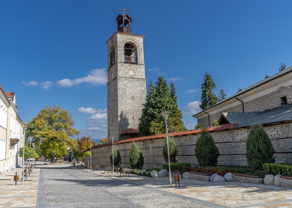 The stone clock tower of the Holy Trinity church in Bansko, a paved square and autumn trees under a deep blue sky