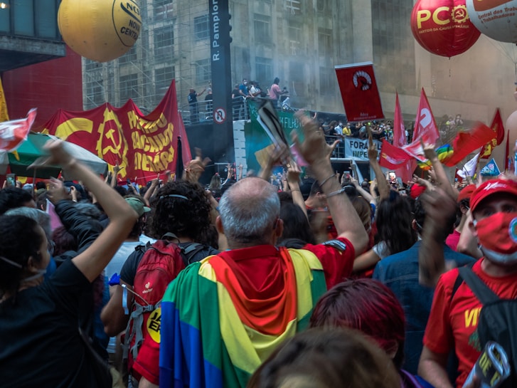 A large crowd of people gathered in an urban setting, holding flags and banners. The scene is energetic, with people cheering and waving colorful flags. Some flags have symbols and writing in another language, suggesting a political or social rally. Bright balloons float above, adding to the festive yet intense atmosphere.