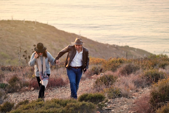 A couple is walking hand in hand on a hilly path surrounded by dry shrubs and grass, with an ocean view in the background. Both are wearing hats and casual outfits suitable for an outdoor adventure.