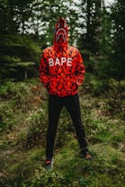A person stands in a forest wearing a vibrant red and black camo-patterned hoodie with the hood pulled up, covering their face. The hoodie features a shark face and the word 'BAPE' across the chest. The background consists of dense green foliage under natural lighting.