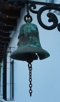 A weathered metal bell with a patina finish hangs from a decorative wrought iron bracket. The bell appears to be aged, with a significant amount of rust and tarnish visible on its surface. A chain hangs from the center of the bell, indicating it is used as a ringing mechanism. In the background, the white wall of a building provides contrast, with shadowed window grills and part of a wooden roof structure visible.