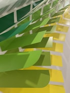 A series of overlapping panels in varying shades of green and yellow are mounted on a wall, creating a dynamic and textured visual pattern. The panels have a rectangular shape with curved edges and are arranged in a cascading manner, producing an eye-catching gradient effect.