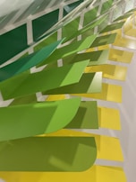A series of overlapping panels in varying shades of green and yellow are mounted on a wall, creating a dynamic and textured visual pattern. The panels have a rectangular shape with curved edges and are arranged in a cascading manner, producing an eye-catching gradient effect.