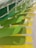 A series of overlapping panels in varying shades of green and yellow are mounted on a wall, creating a dynamic and textured visual pattern. The panels have a rectangular shape with curved edges and are arranged in a cascading manner, producing an eye-catching gradient effect.