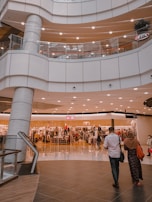 An indoor shopping mall features multiple levels with bright lighting and modern architecture. A clothing store is prominently displayed in the center, with mannequins and racks of clothes visible behind glass windows. Several people are seen walking around, including a man in a white shirt and a woman in a headscarf.