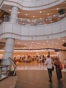 An indoor shopping mall features multiple levels with bright lighting and modern architecture. A clothing store is prominently displayed in the center, with mannequins and racks of clothes visible behind glass windows. Several people are seen walking around, including a man in a white shirt and a woman in a headscarf.