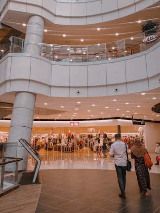 An indoor shopping mall features multiple levels with bright lighting and modern architecture. A clothing store is prominently displayed in the center, with mannequins and racks of clothes visible behind glass windows. Several people are seen walking around, including a man in a white shirt and a woman in a headscarf.