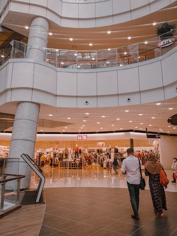 An indoor shopping mall features multiple levels with bright lighting and modern architecture. A clothing store is prominently displayed in the center, with mannequins and racks of clothes visible behind glass windows. Several people are seen walking around, including a man in a white shirt and a woman in a headscarf.