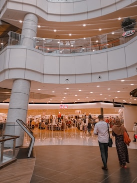 An indoor shopping mall features multiple levels with bright lighting and modern architecture. A clothing store is prominently displayed in the center, with mannequins and racks of clothes visible behind glass windows. Several people are seen walking around, including a man in a white shirt and a woman in a headscarf.