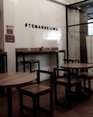A cozy interior setting with wooden tables and chairs, characterized by minimalist black metal frames. The wall features a hashtag sign that reads '#TEMANSEJIWA' in a bold, simple font. The space has a small sign prohibiting smoking, and the overall ambiance is functional and modern, with a mix of wood and metal materials.