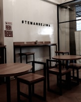 A cozy interior setting with wooden tables and chairs, characterized by minimalist black metal frames. The wall features a hashtag sign that reads '#TEMANSEJIWA' in a bold, simple font. The space has a small sign prohibiting smoking, and the overall ambiance is functional and modern, with a mix of wood and metal materials.