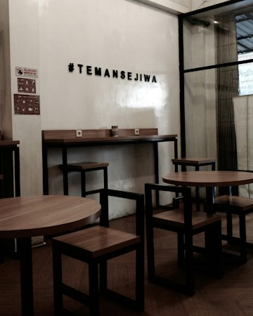 A cozy interior setting with wooden tables and chairs, characterized by minimalist black metal frames. The wall features a hashtag sign that reads '#TEMANSEJIWA' in a bold, simple font. The space has a small sign prohibiting smoking, and the overall ambiance is functional and modern, with a mix of wood and metal materials.