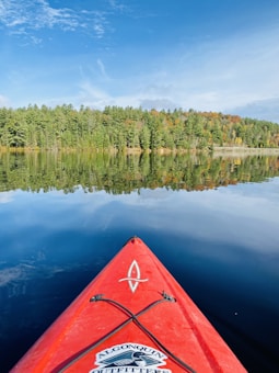 A serene lake reflects a dense tree line of lush green forest under a bright blue sky scattered with a few clouds. The red tip of a kayak is visible in the foreground, suggesting a peaceful paddling experience.