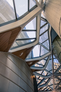 A modern architectural structure featuring a geometric metal framework and glass panels. The design includes triangular and angular shapes with a combination of materials like metal and concrete. The view captures the interplay of shadows and light through the open spaces in the ceiling, while parts of a building exterior are visible through the glass.