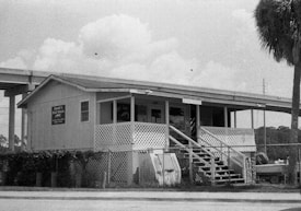 A wooden building with a porch and stairs leading up to the entrance. The structure appears to be elevated with latticework around the base. There is a sign identifying the building as 'Bantee&rsquo;s Bait Shop' and another advertisement for a bait shack. The building is situated near a road, with a palm tree and some vegetation around it. A large overhead structure, possibly a bridge or overpass, is visible in the background along with some power lines.