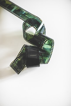 A length of filmstrip is coiled in a loose spiral on a plain, light background. The filmstrip has a glossy appearance with visible perforations along the edges and varying shades of dark, translucent green.