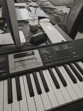 A digital keyboard is placed on a desk, surrounded by a computer monitor, a keyboard, a mouse, and a mason jar with a drink and straw. The setting appears to be an informal workspace with a cozy, lived-in atmosphere, and a plush blanket in the background.