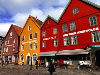 A row of brightly colored, traditional wooden buildings sitting along a cobblestone street. The buildings have large, white-framed windows and some have signage indicating businesses such as a hotel and a nightclub. A few people walk along the street, and the sky is partly cloudy.