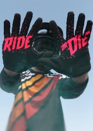 A person wearing a colorful motocross jersey stands with their hands extended forward, displaying black gloves with bold red text that reads 'RIDE OR DIE.' The background is a clear blue sky, and the person is also wearing a helmet.