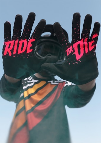 A person wearing a colorful motocross jersey stands with their hands extended forward, displaying black gloves with bold red text that reads 'RIDE OR DIE.' The background is a clear blue sky, and the person is also wearing a helmet.