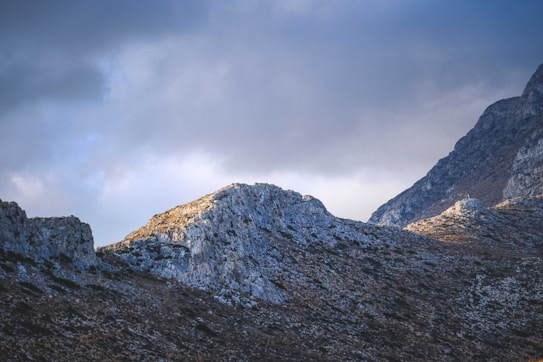 The image features a rugged mountain landscape under a cloudy sky. The peaks are illuminated by soft, filtered sunlight, highlighting the rocky textures and creating contrasting shadows.