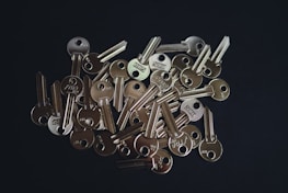 A scattered pile of shiny metallic keys lying on a dark surface. The keys have rounded heads with holes and grooves along their shafts. They reflect light, creating highlights on their surfaces.