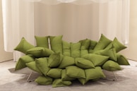 A unique green sofa composed of multiple overlapping cushions, arranged in a modern and artistic fashion. The sofa rests on a metal frame and is placed against a backdrop of white curtains, with a neutral-colored carpet underneath.
