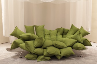 A unique green sofa composed of multiple overlapping cushions, arranged in a modern and artistic fashion. The sofa rests on a metal frame and is placed against a backdrop of white curtains, with a neutral-colored carpet underneath.