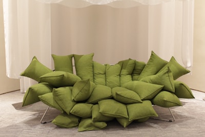 A unique green sofa composed of multiple overlapping cushions, arranged in a modern and artistic fashion. The sofa rests on a metal frame and is placed against a backdrop of white curtains, with a neutral-colored carpet underneath.