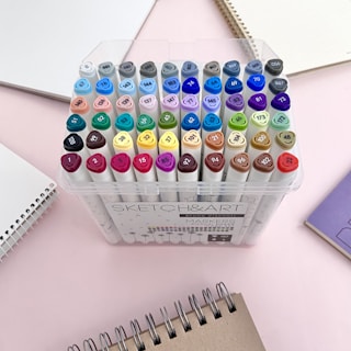 A collection of colorful markers organized in a clear plastic case, surrounded by spiral-bound notebooks on a light pink surface.