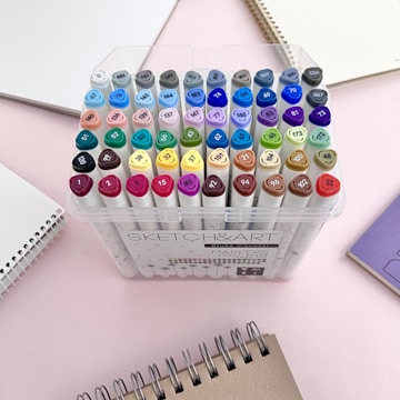A collection of colorful markers organized in a clear plastic case, surrounded by spiral-bound notebooks on a light pink surface.