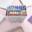 A collection of colorful markers organized in a clear plastic case, surrounded by spiral-bound notebooks on a light pink surface.