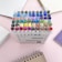 A collection of colorful markers organized in a clear plastic case, surrounded by spiral-bound notebooks on a light pink surface.