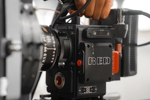 A high-end video camera with a prominent lens and the brand name 'RED' visible on the side. Various buttons and adjustment knobs are seen, along with part of a hand holding the camera.